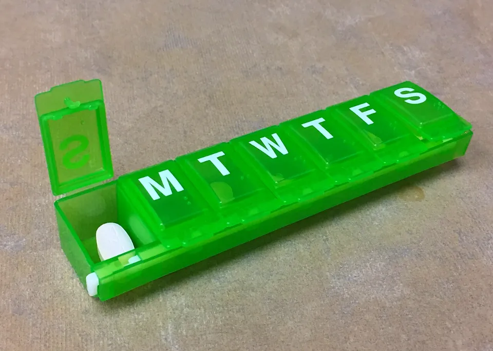 A green seven-day pill organizer with one compartment open and a white tablet inside