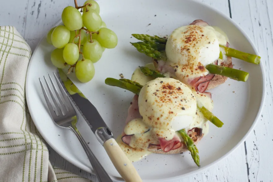 A white plate with poached eggs, ham, asparagus and grapes, served on a wooden table