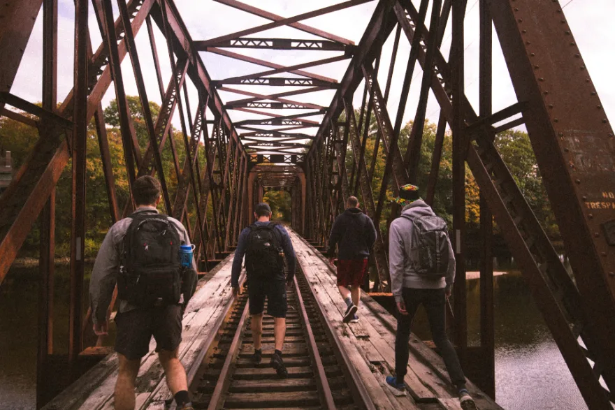 Four adults walking outdoors on a bridge in daylight, seen from behind
